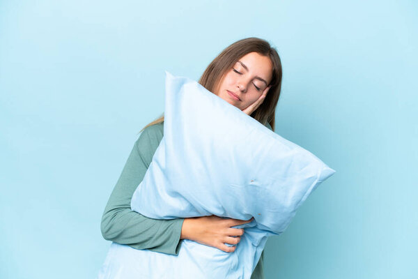 Young beautiful woman isolated on blue background in pajamas and holding a pillow while sleeping