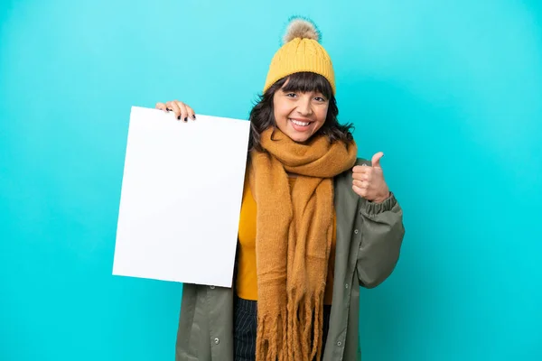 stock image Young latin woman wearing winter jacket isolated on blue background holding an empty placard with thumb up