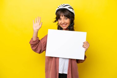Genç bisikletçi Latin kadın, sarı arka planda izole edilmiş, elinde boş bir levha, selam veriyor.