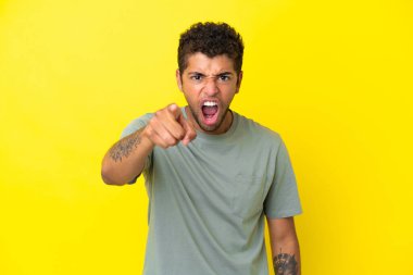 Genç yakışıklı Brezilyalı adam sarı arka planda izole edilmiş hayal kırıklığına uğramış ve ön tarafı işaret ediyor.