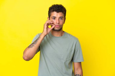 Sarı arka planda izole edilmiş yakışıklı Brezilyalı genç bir adam sessizlik işareti gösteriyor.