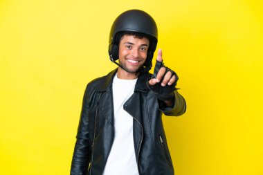 Sarı arka planda izole edilmiş motosiklet kasklı genç Brezilyalı adam parmağını kaldırıyor ve gösteriyor.