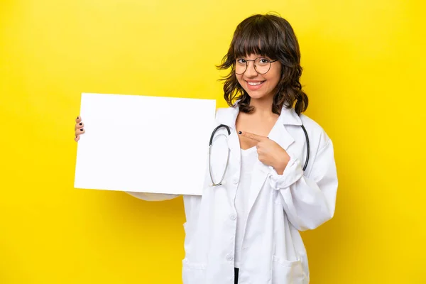 年轻的医生拉汀女士 在黄色的背景上被隔离着 手里拿着一张空空的标语牌 脸上带着快乐的表情 指着它 — 图库照片