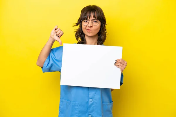 一个年轻的护士医生女人 被隔离在黄色背景下 手里拿着一个空标语牌 做着坏的信号 — 图库照片