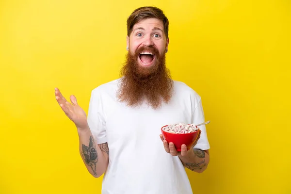 Redhead man with beard eating a bowl of cereals isolated on yellow background with shocked facial expression