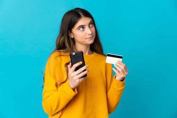 Young caucasian woman isolated on blue background buying with the mobile with a credit card while thinking