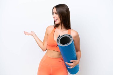 Genç ve güzel spor kadın yoga derslerine giderken beyaz arka planda bir minder tutuyor ve sürpriz bir ifadeyle yan yana bakıyor.