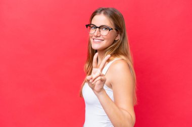 Kırmızı arka planda izole edilmiş güzel bir kadın gülümsüyor ve zafer işareti gösteriyor.