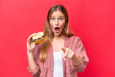 Elinde hamburger tutan beyaz bir kadın kırmızı arka planda izole edilmiş, şaşırmış ve ön tarafı işaret ediyor.