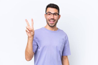 Beyaz arka planda izole edilmiş genç beyaz adam gülümsüyor ve zafer işareti gösteriyor.