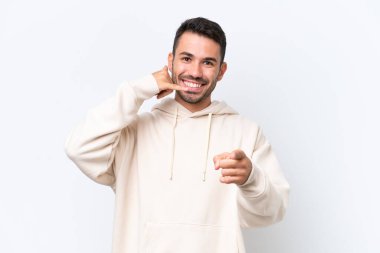 Beyaz arka planda izole edilmiş genç beyaz bir adam telefon jesti yapıyor ve ön tarafı işaret ediyor.