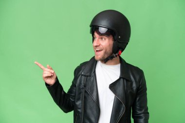 Yeşil krom arka planda izole edilmiş motorsiklet kasklı genç beyaz bir adam. Parmakla yan tarafı gösteriyor ve bir ürün sunuyor.