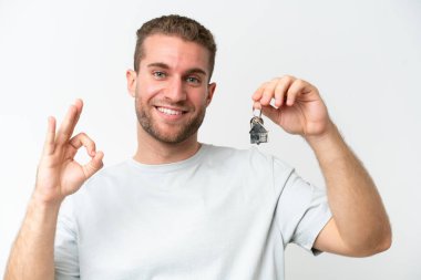 Young caucasian man holding home keys isolated on white background smiling and showing victory sign