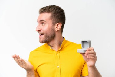 Elinde nişan yüzüğü olan genç bir adam beyaz arka planda, şaşırmış bir yüz ifadesiyle izole edilmiş.