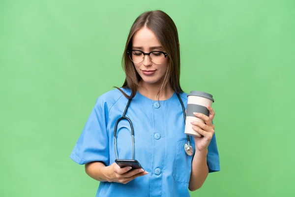 stock image Young nurse caucasian woman over isolated background holding coffee to take away and a mobile