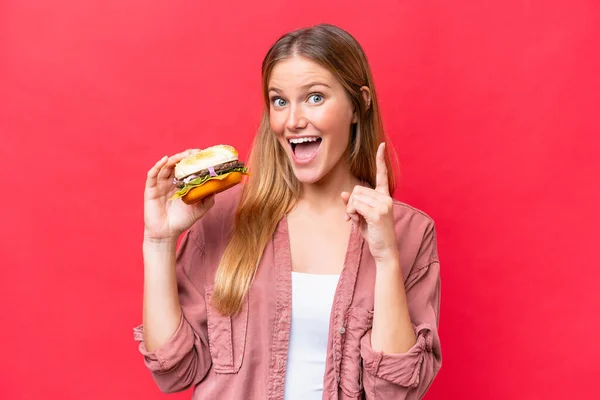 Mladá Běloška Drží Burger Izolovaný Červeném Pozadí Ukazuje Skvělý Nápad — Stock fotografie