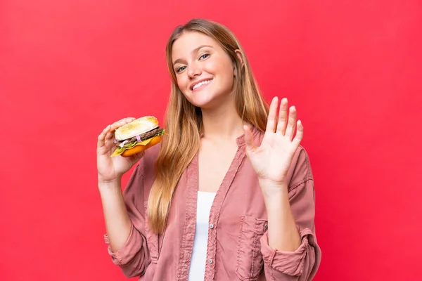 Młoda Kaukaska Kobieta Trzyma Hamburgera Odizolowany Czerwonym Tle Salutując Ręka — Zdjęcie stockowe