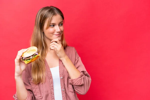 Młoda Kaukaska Kobieta Trzyma Hamburgera Odizolowany Czerwonym Tle Myśląc Pomysł — Zdjęcie stockowe