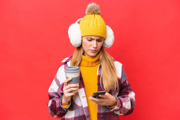 Молодая красивая женщина в зимних муфтах изолированы на красном фоне держа кофе, чтобы забрать и мобильный
