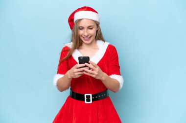 Ana baba gibi giyinmiş genç sarışın kadın mavi arka planda izole edilmiş cep telefonuyla bir mesaj gönderiyor.