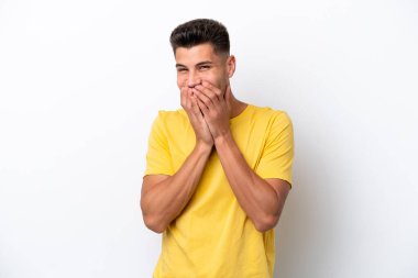 Beyaz arka planda izole edilmiş genç beyaz bir adam mutlu ve gülümseyen elleriyle ağzını kapatıyor.