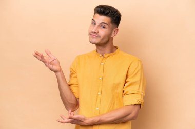 Beyazlı genç bir adam bej arka planda izole edilmiş. Davet etmek için ellerini yana uzatıyor.