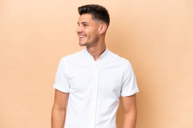 Beyazlı genç bir adam bej arka planda izole edilmiş, yana bakıyor ve gülümsüyor.