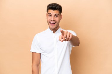 Beyazlı genç adam bej arka planda izole edilmiş şaşırmış ve ön tarafı işaret ediyor.