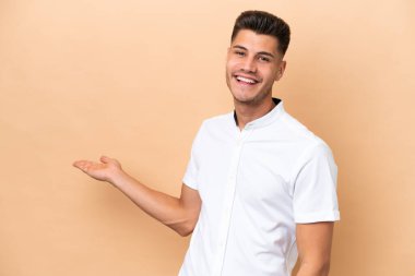 Beyazlı genç bir adam bej arka planda izole edilmiş. Davet etmek için ellerini yana uzatıyor.