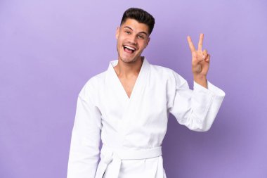 Mor arka planda karate yapan genç beyaz adam gülümsüyor ve zafer işareti gösteriyor.