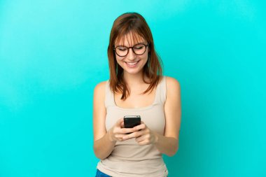 Kızıl saçlı kız mavi arka planda izole edilmiş. Cep telefonuyla mesaj gönderiyor.