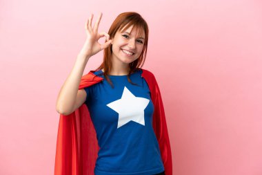 Süper kahraman kızıl saçlı kız pembe arka planda izole edilmiş. Parmaklarıyla tamam işareti gösteriyor.