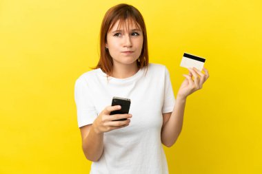 Redhead girl isolated on yellow background buying with the mobile with a credit card while thinking