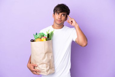 Elinde alışveriş çantası tutan genç bir adam mor arka planda izole edilmiş kuşkuları ve düşünceleri var.