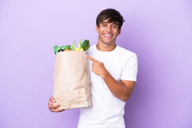 Elinde mor arka planda izole edilmiş bir alışveriş çantası tutan genç adam bir ürünü sunmak için yan tarafı işaret ediyor.