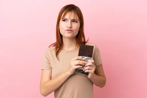 Roodharige Meisje Geïsoleerd Roze Achtergrond Nemen Van Een Chocolade Tablet — Stockfoto