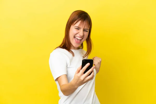Rotschopfiges Mädchen Isoliert Auf Gelbem Hintergrund Mit Handy Und Siegesgeste — Stockfoto