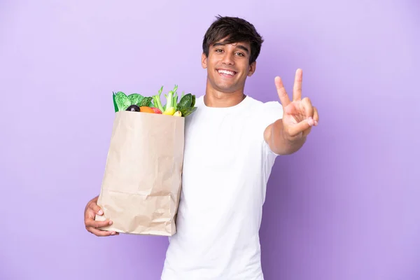 Νεαρός Άνδρας Κρατώντας Μια Τσάντα Ψώνια Απομονωμένη Μωβ Φόντο Χαμογελώντας — Φωτογραφία Αρχείου