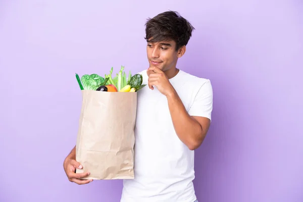 Young man holding a grocery shopping bag isolated on purple background looking to the side and smiling