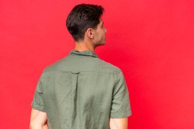 Beyaz tenli yakışıklı adam kırmızı arka planda izole edilmiş ve arkasına bakıyor.