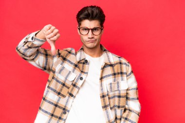 Genç, beyaz, yakışıklı bir adam kırmızı arka planda izole edilmiş, olumsuz bir ifadeyle baş parmağını aşağı gösteriyor.