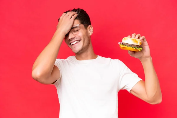 Młody Biały Mężczyzna Trzymając Hamburgera Izolowany Czerwonym Tle Zdał Sobie — Zdjęcie stockowe