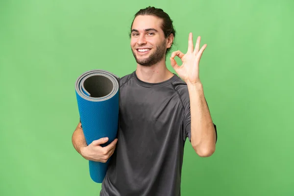 年轻的运动员一边去上瑜伽课 一边拿着垫子盖住孤立的背景 用手指显示出好的手势 — 图库照片