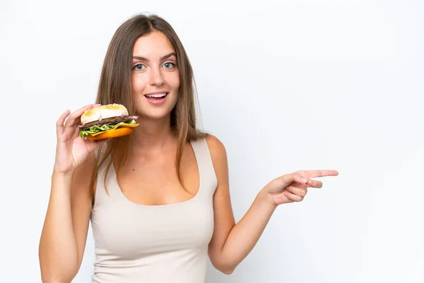 Młoda Ładna Kobieta Trzyma Hamburgera Odizolowany Białym Tle Zaskoczony Wskazując — Zdjęcie stockowe