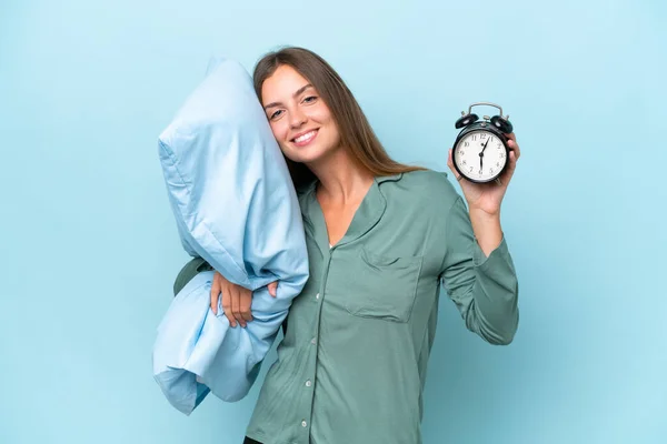 穿着睡衣 穿着蓝色背景的年轻漂亮的女人 带着欢乐的表情 拿着钟和枕头 — 图库照片