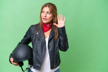 Arka planda motosiklet kaskı olan orta yaşlı beyaz bir kadın kulağına bir şey dayayarak bir şeyler dinliyor.