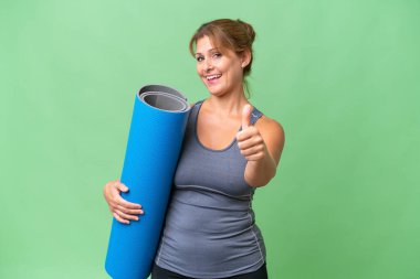 Orta yaşlı sporcu bir kadın yoga derslerine gidiyor. Elinde paspas tutarken. Başparmakları havada.