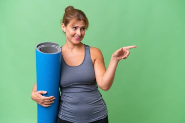 Orta yaşlı sporcu bir kadın yoga derslerine giderken arka planda izole edilmiş bir paspas tutuyor, parmağını yana uzatıyor ve bir ürün sunuyor.