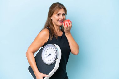 Orta yaşlı beyaz kadın, mavi arka planda, tartı makinesi ve elmayla izole edilmiş.