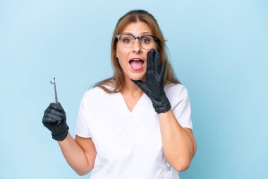 Orta yaşlı dişçi, mavi arka plan üzerinde aletler tutuyor ve ağzı açık bağırıyor.